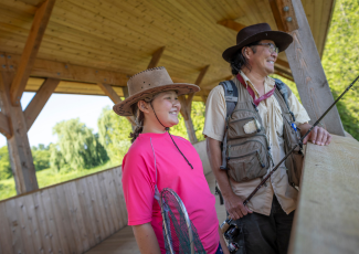 A father and daughter hold fishing rods and a net while standing on a bridge and smiling as they look at the scenery.